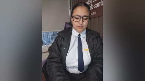 Video of a Latina woman with light skin, wearing glasses, a white shirt, navy tie, and black blazer, sitting in a government office with blue and white patterned wallpaper and a brown sign.