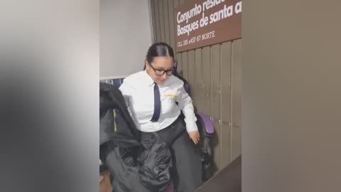 Media: Video of a woman with dark hair, glasses, and black coat, unzipping it, standing in a corridor with a sign reading \"Correos de Santa Fe.\