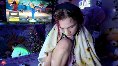 Video of a young, fair-skinned woman with wet, shoulder-length brown hair, wearing a yellow towel, seated in a cluttered, dimly lit room with gaming console and plush toys.