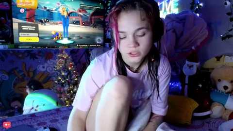 Video of a young, fair-skinned girl with wet, shoulder-length hair, wearing a pink T-shirt and white shorts, sitting in a cluttered room with a TV showing Mario Kart, stuffed animals, and a Christmas tree.