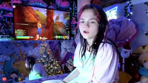 Video of a young girl playing a video game, wet hair, wearing a white shirt, surrounded by colorful, cartoon-themed decorations, including a Christmas tree, in a cozy, playful room.