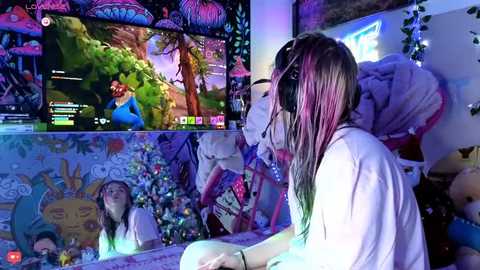Video of a young woman with long, multicolored hair wearing headphones, sitting in a colorful, eclectic room with a large TV screen showing a video game. The room is decorated with vibrant wall art, plush toys, and fairy lights.