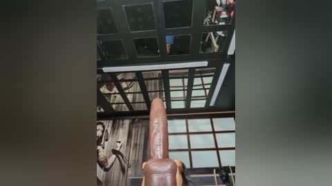 Video of a modern, open-plan office with a tall, realistic wooden dildo sculpture standing prominently in the center. The background features a grid of glass panels and black metal frames.