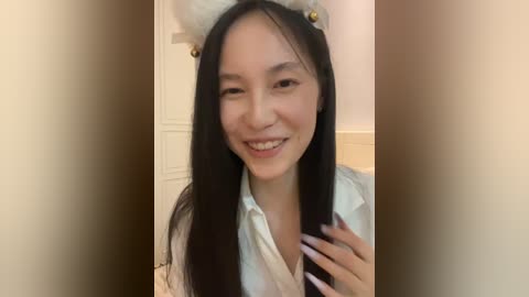 Video of an East Asian woman with long black hair, wearing a white shirt, and a fluffy white cat headband with gold bells. She smiles widely with braces, indoors, with a white door in the background.