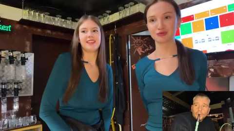 Video of a smiling, brunette woman in a teal V-neck sweater, standing in a bar with shelves of liquor bottles and a colorful digital clock on the wall.