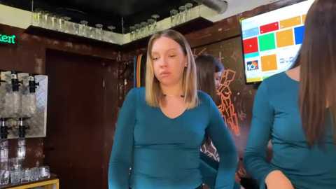 Video of a blonde woman with shoulder-length hair, wearing a teal long-sleeve top, standing in a dimly lit bar with shelves of liquor bottles, a colorful television screen, and another woman in the background.