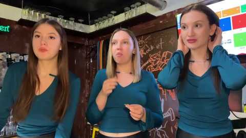 Video of three young women in teal V-neck shirts, lipsyncing and dancing in a dimly-lit, graffiti-covered bar with shelves of alcohol.