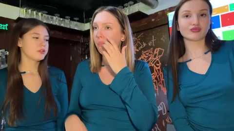 Video of three young women with straight, long brown hair, wearing teal long-sleeved shirts, seated indoors, one with hand on cheek, another blowing kiss, and third looking down, against a colorful, graffiti-covered wall.