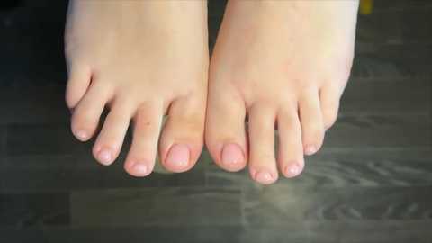 A close-up video of two bare feet with light skin tones, clean nails, and a soft, smooth texture. The feet are positioned on a dark wooden floor with a subtle grain pattern, creating a minimalistic and clean aesthetic.