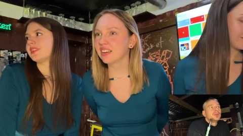 Video of three young women with long hair, wearing blue tops, standing in a dimly lit bar with graffiti on the wall and a large TV screen displaying a colorful logo.