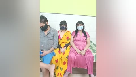 Video of three people in a waiting room. Two women in pink and yellow saris with masks, one man in a gray shirt and blue shorts.