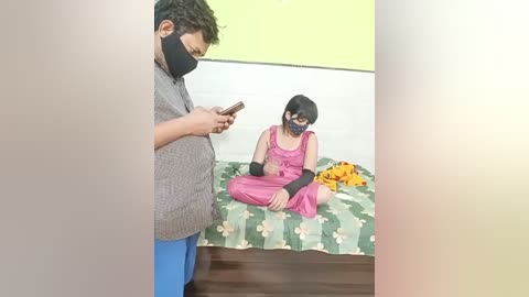 A video shows a South Asian man in a grey shirt and blue pants, standing in a room with a masked woman in pink pajamas sitting on a bed with floral bedding.