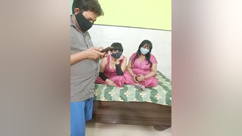 Video of a man with short hair in a gray shirt, blue pants, black mask, cutting a woman's hair in a small room with a green floral bedspread and yellow wall.