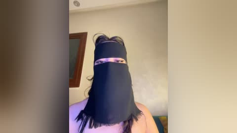 A video of a person wearing a black niqab with long fringe, standing indoors against a beige wall with a framed picture. The person's face is obscured, revealing only their eyes.