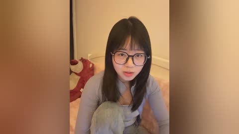 Video of a young Asian woman with straight black hair and glasses, wearing a light blue shirt, sitting on a beige carpet in a narrow hallway, with red slippers and a red bag in the background.