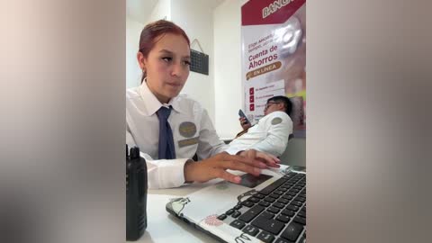Video of a young woman in a white uniform with a blue tie, using a laptop in a clinic. Patient lying on a bed in the background.