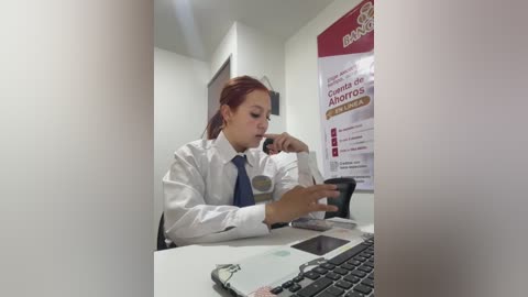 A video of a young woman with light skin, red hair, and a white shirt, working on a computer in a brightly lit office.
