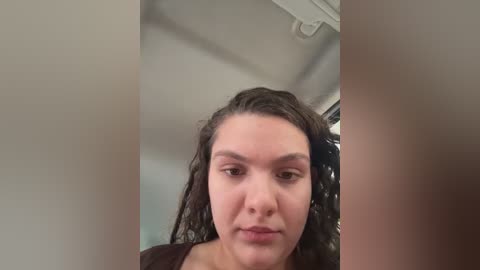 A video of a young woman with curly dark hair, light skin, and neutral expression, partially obscured by two vertical objects. The background shows an indoor setting with white walls and a ceiling light.