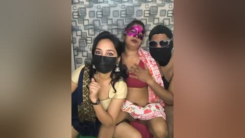 A video of three South Asian women in masks, wearing colorful traditional outfits, against a geometric-patterned wall.