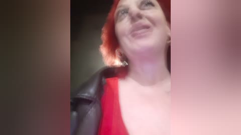 Video of a woman with short, fiery red hair, wearing a red top and black leather jacket, smiling, with a blurred, dimly lit background.