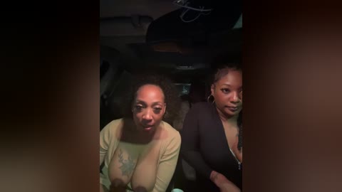 Video of two Black women in a dimly-lit car, one with a large tattoo on her arm and the other wearing a black top.