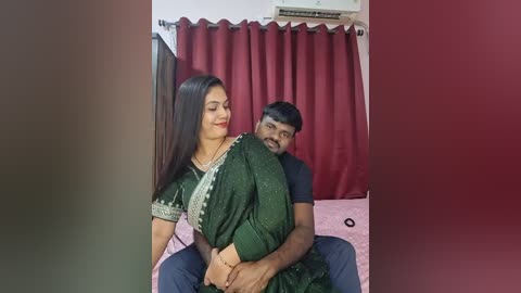 A video of a South Asian couple in traditional attire, the woman in a green sari and the man in a black shirt, sitting closely together on a bed with red curtains and a pink blanket.