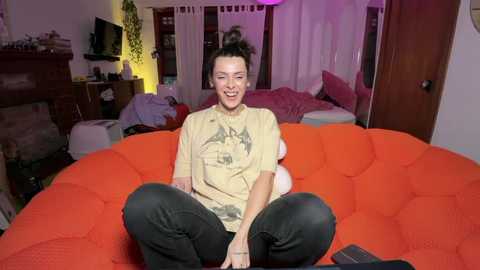 Media: Video of a smiling young woman with short dark hair, wearing a beige graphic t-shirt and black pants, sitting on a bright orange beanbag chair in a cluttered, dimly-lit bedroom with purple and white curtains.