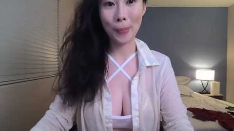 Media: Video of an Asian woman with long, wavy black hair, wearing a sheer white blouse and white bra, in a modern bedroom with a bed, lamp, and window blinds.