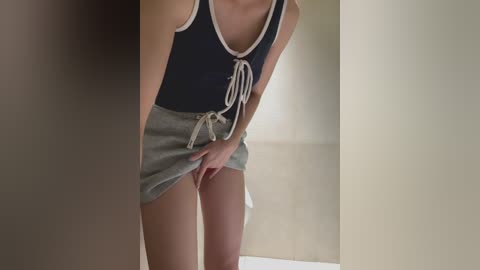 Video of a slender, fair-skinned woman with a small bust, wearing a dark blue tank top with white trim and a grey skirt. She is indoors, with a blurred background.