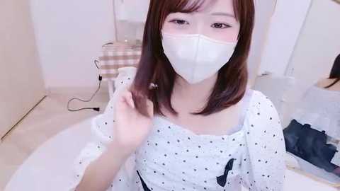 Video of an Asian woman with light skin and shoulder-length brown hair, wearing a white mask and polka-dot dress, sitting on a bed, adjusting her hair.