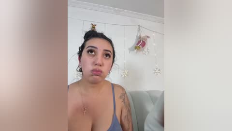 Video of a Latina woman with medium skin tone, dark hair in a bun, wearing a light blue spaghetti strap top, standing indoors with white walls and festive decorations, making a pouty face.