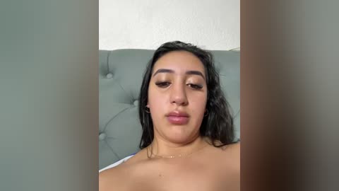 Video of a young woman with medium brown skin, long black hair, and full lips, wearing no shirt, lying on a gray tufted sofa with a white wall in the background.