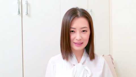 Video of an East Asian woman with straight, shoulder-length brown hair, wearing a white blouse, standing against a white wardrobe with silver handles.