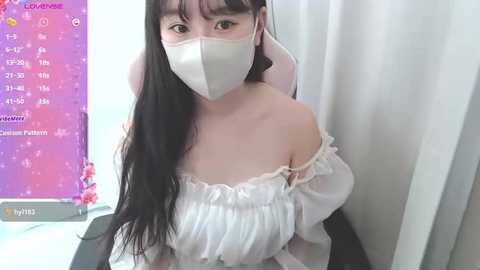 A video of an Asian woman with long black hair, wearing a white off-shoulder dress, face mask, and headphones, seated in a dimly lit room with a pink and purple computer screen.