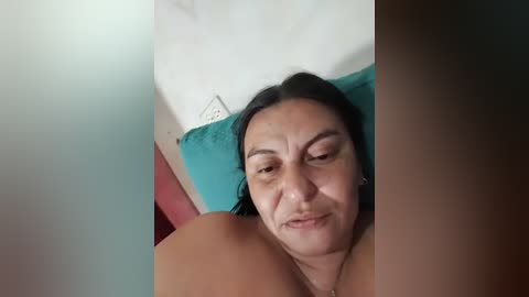 Video of a middle-aged woman with medium skin tone, dark hair, and a neutral expression, lying on a teal pillow. The background features a white wall and a red object.