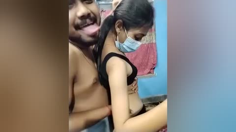 Media: Video of a topless South Asian man with a light brown complexion and a medium build, leaning over a partially clothed woman with a dark complexion, who wears a mask and black bra, in a room with blue walls and a red towel hanging.