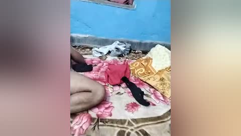 Video of a cluttered room with a blue wall and tiled floor. A woman in a pink floral dress sits on a patterned rug, surrounded by discarded clothing and bedding.