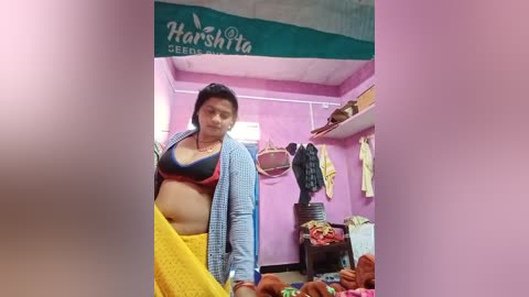 Video of a pregnant woman in a small, cluttered room with purple walls and green curtains. She wears a yellow sari, blue cardigan, and black bra, holding a baby in her arms.