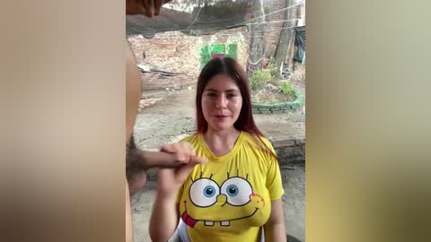 A video shows a young woman with long, straight brown hair, wearing a yellow SpongeBob T-shirt, in a construction site with exposed wires, green plastic barriers, and dirt. She holds a man's hand.