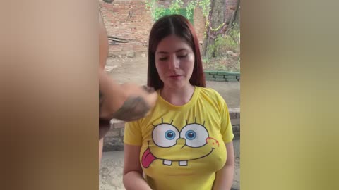 Video of a young woman with fair skin and long brown hair, wearing a yellow SpongeBob shirt, smiling while a tattooed man's hand is in the frame.