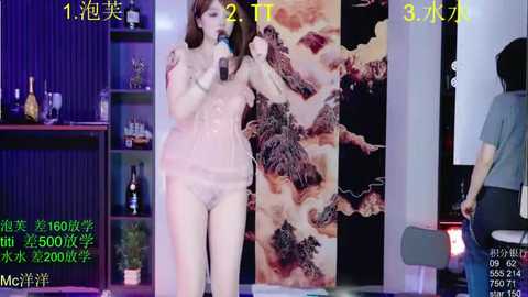 A video captures a nude Asian woman with fair skin, medium breasts, and long brown hair, standing in front of a modern room with a large, artistic painting on the wall.