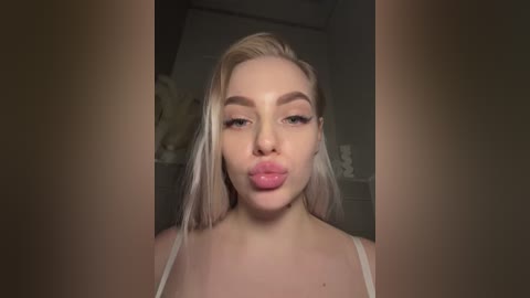 Video of a young woman with platinum blonde hair, wearing a white spaghetti-strap top, puckering her lips in a playful manner. She has a fair complexion and a slight, confident smile.
