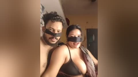 Video of a couple with blindfolds and lingerie, standing close in a dimly-lit room with beige walls and a door visible in the background.