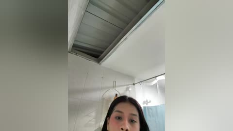 A video of an Asian woman with long black hair, wearing a white shirt, standing in a dimly lit, small bathroom with a ceiling vent and shower curtain.