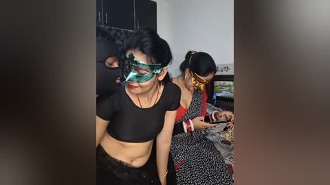 Video of three women in a domestic setting; two wear black masks, one with a green mask, and all wear traditional Indian attire.