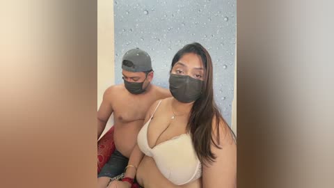 Video of a topless man and woman wearing face masks, both with medium skin tones, in a bathroom with a textured gray wall. The woman is in a beige bra, while the man is shirtless, wearing a baseball cap.