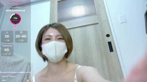 Video of a young East Asian woman with short brown hair, wearing a white face mask, light beige bralette, and a light wooden door background, with a temperature gauge overlay showing 20.6\u00b0C.