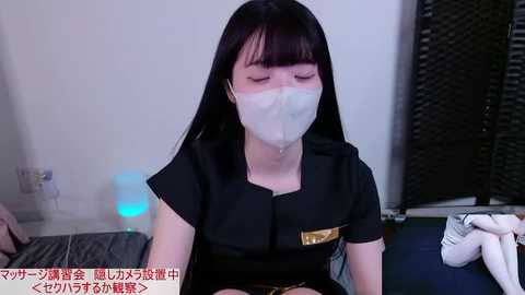 Video of a young Asian woman with long black hair, wearing a black outfit and white face mask, sitting on a bed in a dimly lit room, with Japanese text at the bottom.