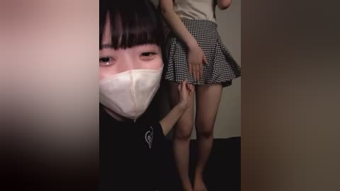 Media: Video of an East Asian woman in a black mask, black shirt, and white face shield, standing beside a young girl in a checkered skirt, holding her hand.
