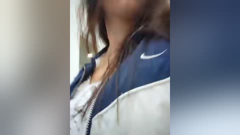 Video of a close-up of a person's neck and chest, wearing a white and blue Nike jacket with a visible zipper, and a white lace bra peeking through. The background is blurred, featuring a gradient of blue and beige tones.